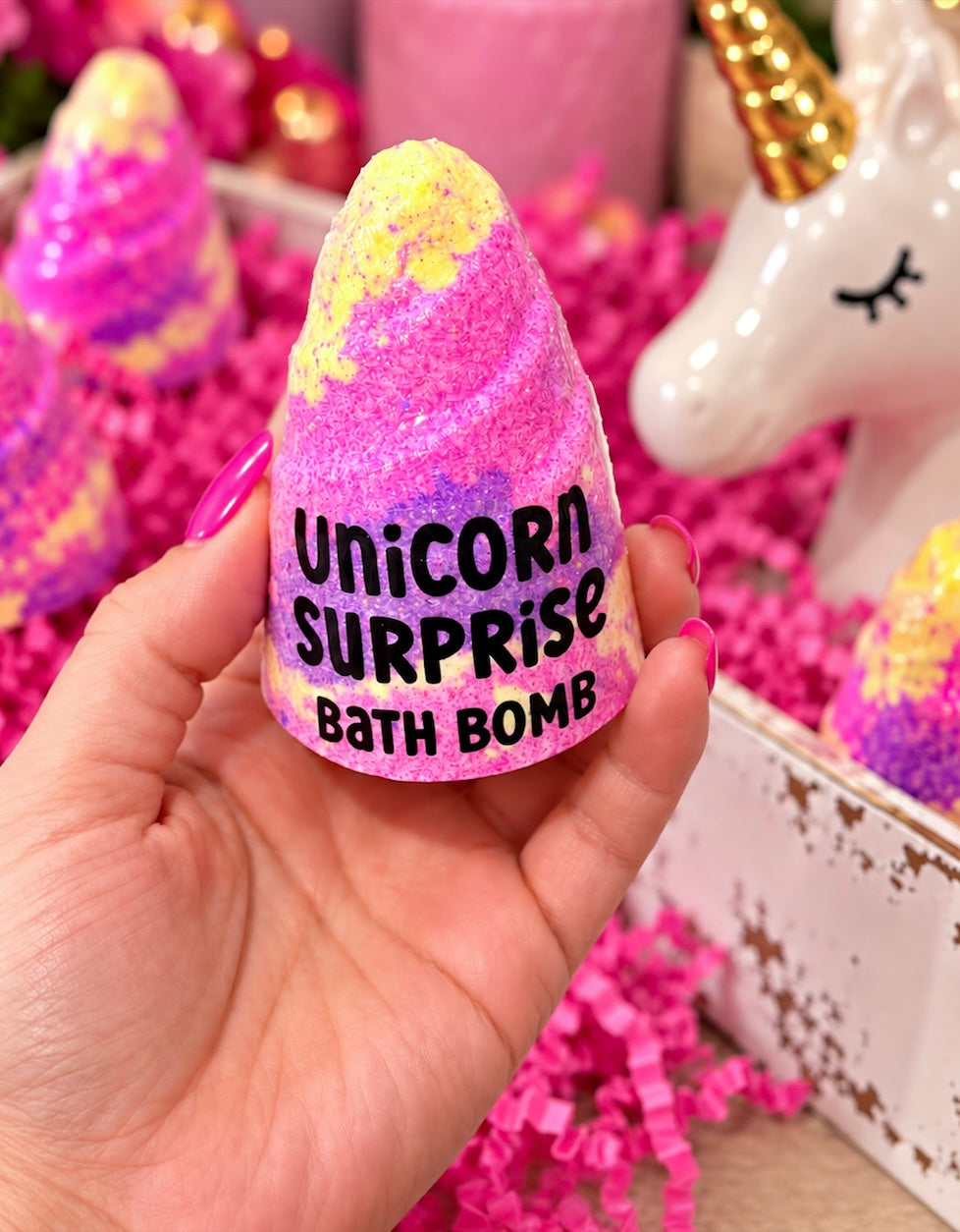 A hand holds a pink and yellow glitter bath bomb shaped like a unicorn head, labeled 'Unicorn Surprise Bath Bomb'. The background is filled with pink shredded paper, other colorful bath bombs, and a white unicorn figurine.