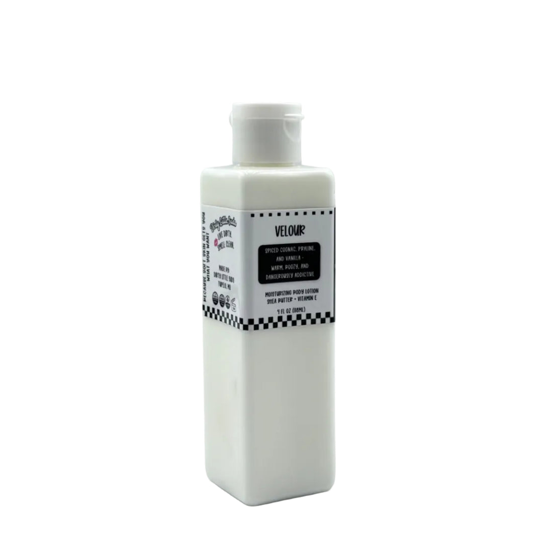 Dirty Little Suds Velour body lotion in a white rectangular bottle with flip-top cap and black label, featuring a warm spiced cognac, praline, and vanilla scent.
